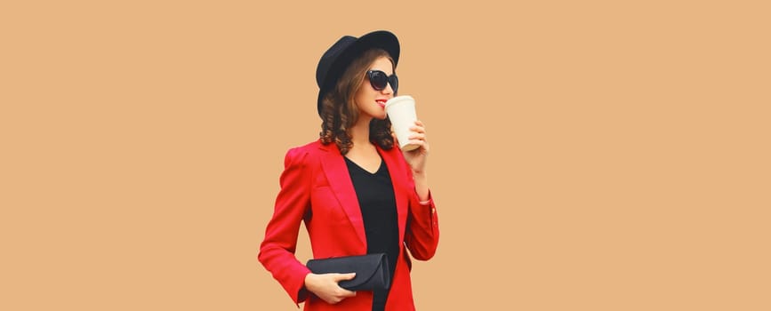 Stylish elegant woman posing in business suit, red blazer jacket, black round hat with handbag clutch looking away isolated on brown studio background