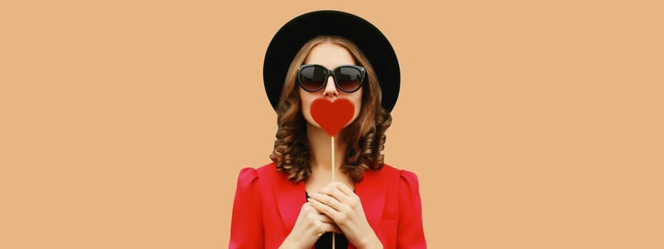 Beautiful stylish woman blowing kiss with sweet red heart shaped lollipop on stick in black round hat on brown background
