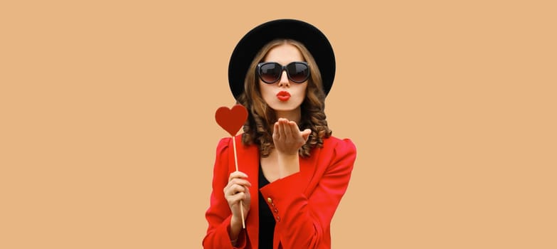 Beautiful stylish woman blowing kiss with sweet red heart shaped lollipop on stick in black round hat on brown background