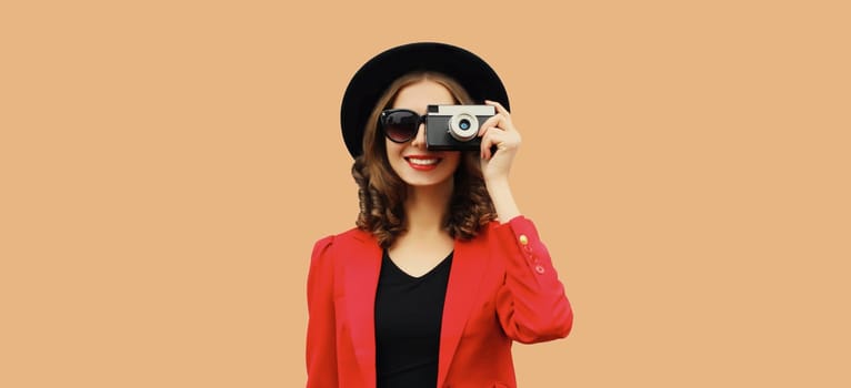 Beautiful stylish young woman photographer with film camera taking picture in black round hat, red blazer jacket isolated on brown studio background