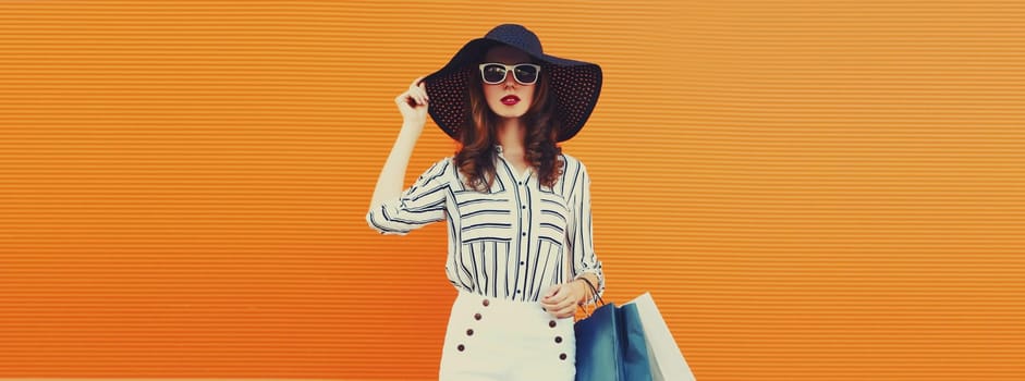 Stylish beautiful young woman model with shopping bags in summer black straw hat, striped dress on orange background