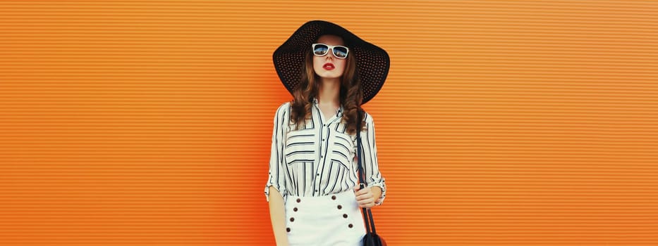 Portrait of beautiful young woman posing wearing a white striped shirt, summer black round hat on orange background