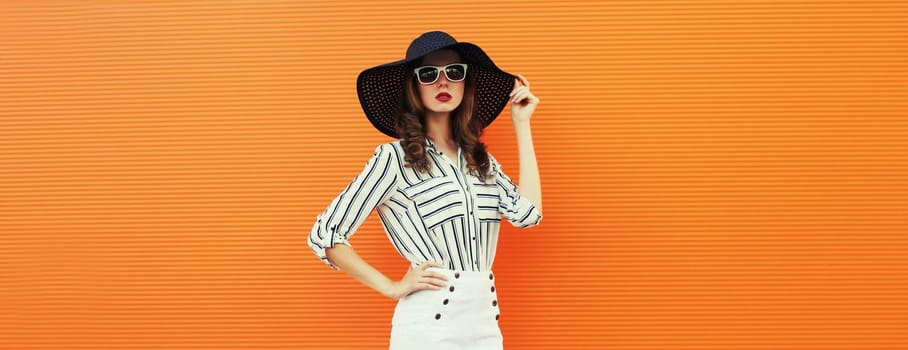 Portrait of beautiful young woman posing wearing a white striped shirt, summer black round hat on orange background