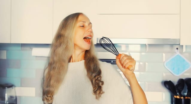 Happy cheerful middle aged woman singing with whisk having fun in the kitchen at home in the morning