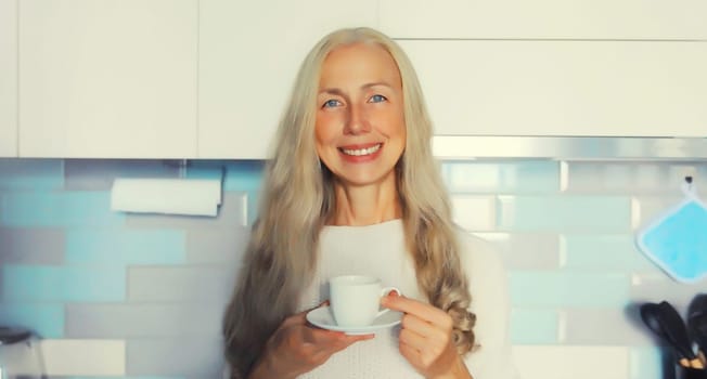 Portrait of happy smiling middle-aged gray-haired woman enjoying hot tasty cup of coffee or tea in the kitchen in early morning at home