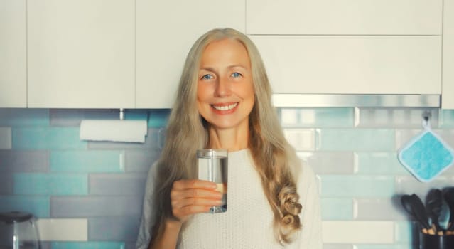 Portrait of happy smiling middle-aged gray-haired woman holds glass with fresh clean water in the kitchen in early morning at home