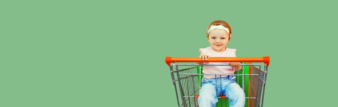 Happy baby with shopping bags sitting in trolley cart on green background, blank copy space for advertising text