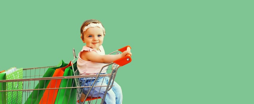 Happy baby with shopping bags sitting in trolley cart on green background, blank copy space for advertising text