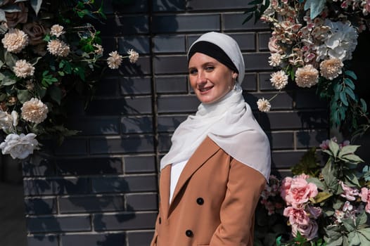 Portrait of a young caucasian woman dressed in a hijab near a wall with flowers