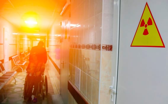 A person in a wheelchair pushes through a hospital corridor with a bright light illuminating the way. A yellow warning sign with a red radiation symbol is prominently displayed on the wall, highlighting the stark contrast between danger and hope.