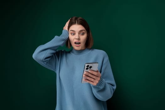 A woman wearing a blue sweater is holding a cell phone in her hand. She appears to be looking at the screen intently, possibly texting or browsing. The background is nondescript.