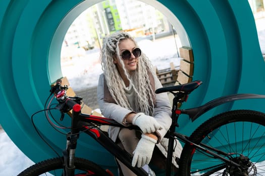 A woman sits on a stationary bike positioned in front of a large circular window. She appears relaxed, looking out through the glass. The surroundings are unseen, focusing on the interaction between the woman and the unique window.