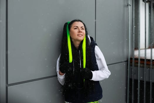 A woman with vibrant green hair is standing against a neutral-colored wall. She appears confident and stylish in her unique hairstyle, contrasting with the plain background.