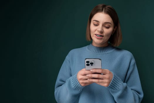 A woman wearing a blue sweater is engrossed in her cell phone screen, her attention focused on the device in her hands as she stands in a room illuminated by natural light.