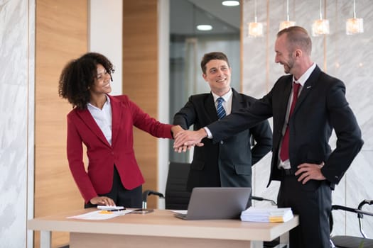 Diverse business team of three people, including a woman and two men, joining hands in a gesture of unity at an office desk with a laptop and paperwork, bright modern office setting