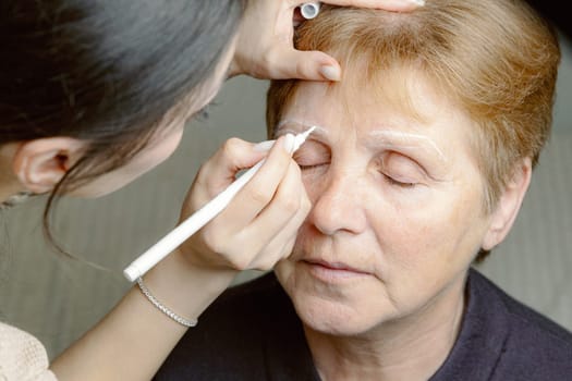 One young Caucasian girl cosmetologist draws a contour with a white marker on the right eyebrow of an elderly beautiful woman with her eyes closed, sitting in a home beauty salon, top side close-up view.
