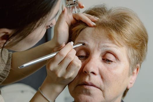 One young Caucasian brunette young girl cosmetologist carefully applies dark brown paint with a brush on the right eyebrow with a white outline to an elderly woman with closed eyes sitting in a home beauty salon, close-up side view.