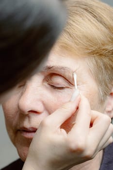 One young Caucasian young brunette girl cosmetologist carefully removes with a cotton stick the remaining paint from the left eyebrow of an elderly woman with closed eyes, looking to the side using a cleanser, in a home beauty salon, side view close-up.