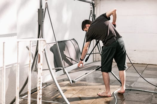 Portrait of a young brunette Caucasian guy in black clothes barefoot washing a garden swing in the backyard of his house with a pressure washer, close-up view from the side.