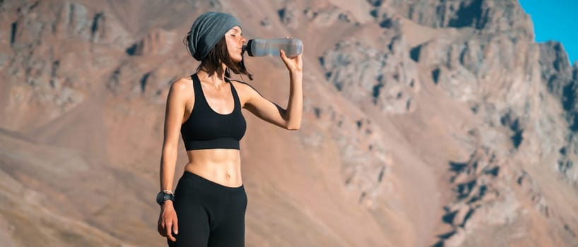 A young girl of athletic build drinks water, isotonic from a bottle during training on trail running in the snow-capped mountains. Runner is making workout outdoor.