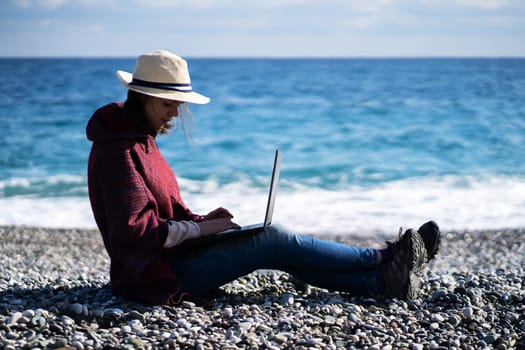 Young girl in a light hat and casual clothes sits on the beach by the sea with a laptop on a sunny day, works, studies, buys tickets during a trip, a woman rests on vacation and types on the keyboard.