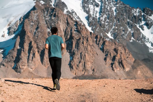 A young man runs along a trail in the snow-capped mountains, doing outdoor workout. The runner warms up, prepares for the race, leads a healthy active lifestyle.