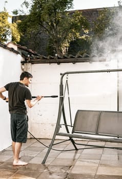 Portrait of a young brunette Caucasian guy in black clothes barefoot from the back washing a garden swing in the backyard of his house with a jet of water under pressure with a kercher, close-up view from the side.