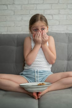 A cute little girl covered in chocolate eats cookies while sitting on the sofa. Vertical photo