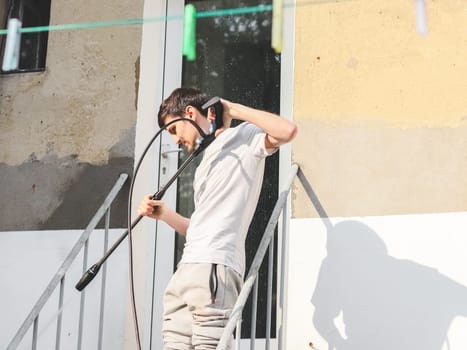 A caucasian guy in gray sports and a T-shirt from the back washes a window with a karcher with water under pressure while standing on a metal staircase in the backyard of his house, side view close-up.The concept of washing the yard,holiday preparation,at home,cleaning services.