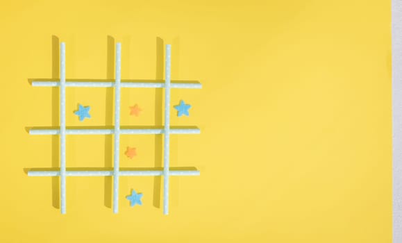 Blue straws with white polka dots lie in a lattice, and inside the squares, felt stars lie on the left on a yellow background with copy space on the right, flat lay, close-up. Concept summer board games.