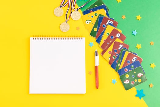 Old,dirty,colorful ligretto game cards,three winner medals,blank notepad,pen and felt stars lie on the right on a green-yellow background with copy space in the left,flat lay close-up.Summer board games concept.