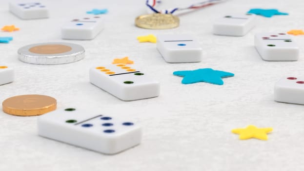 White dominoes with colorful dots, winner medal and chocolate coins lie on a white cement background, close-up side view. Concept summer board game.