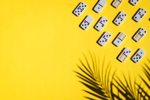 White dominoes with colorful dots lie on the right on a yellow background with a shadow of a palm tree branch with copy space on the left,flat lay close-up.Summer board game concept.