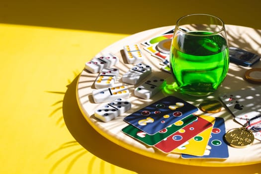 White dominoes, playing cards, uno, ligretto, chocolate coins, winner's medal and green drink in a glass on a round wooden spinning board on the right on a yellow background with a shadow of a palm tree branch and copy space on the left, close-up side view. Concept of a summer board game.