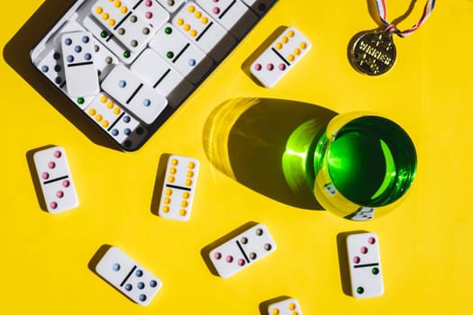 White domino, winner medal and green drink in a glass on yellow background, closeup flat lay. Summer board game concept, banner.