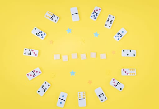 White dominoes with colored dots and empty wooden cubes are arranged in a circle on a yellow background with copy space in the center, closeup flat lay. Summer board game concept.