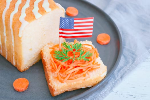 A delicious cake with sugar icing, fresh carrots, American paper flag and chopped pieces in a plate lies on a wooden table, close-up side view. Carrot cake national day concept.