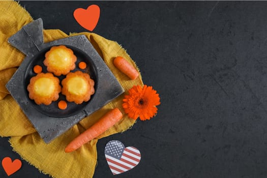 Three carrot muffins in a plate on a gray cutting board on a yellow kitchen napkin with an American flag, red paper hearts lie on the left on a black background with copy space on the right, flat lay close-up. The concept of homemade cakes and carrot cake day.