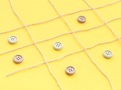 Pale lilac woolen threads lie in a lattice, and inside the squares there are buttons on a yellow background, side view, close-up. Concept summer board games.