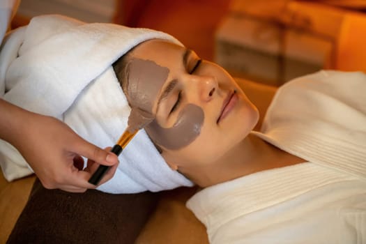 Serene ambiance of spa salon, woman customer indulges in rejuvenating with charcoal face cream massage with warm lighting candle. Facial skin treatment and beauty care concept. Quiescent