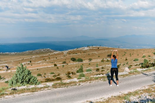 A determined female athlete stretches her muscles after a strenuous run through rugged mountain terrain, surrounded by breathtaking rocky landscapes.