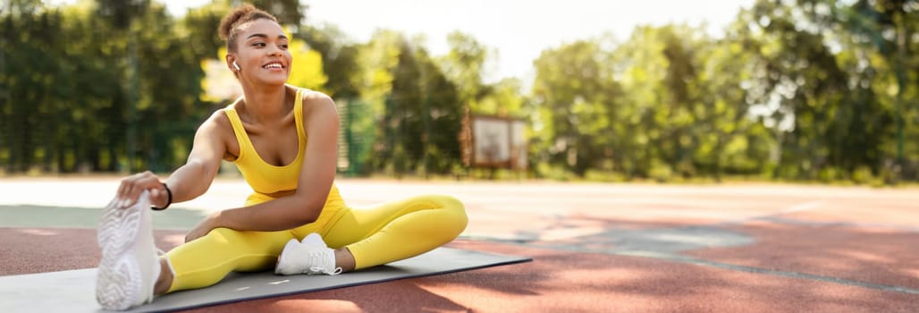 Sporty Lifestyle Concept. Smiling African American lady in yellow top, leggings and white sneakers stretching back, arms and leg muscles sitting on yoga mat outside, touching feet, copy space