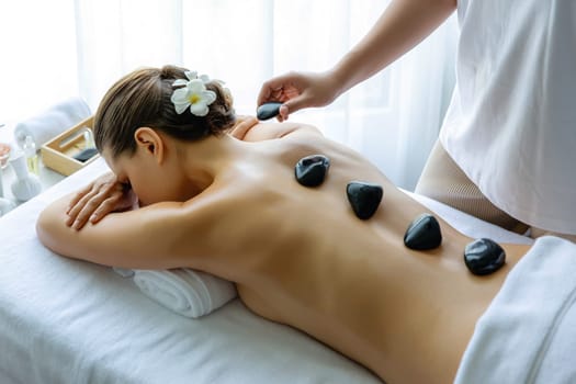 Hot stone massage at spa salon in luxury resort with day light serenity ambient, blissful woman customer enjoying spa basalt stone massage glide over body with soothing warmth. Quiescent