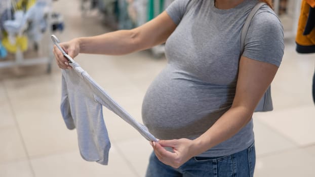 Caucasian pregnant woman chooses baby clothes in a store. Faceless expectant mother in the 3rd trimester