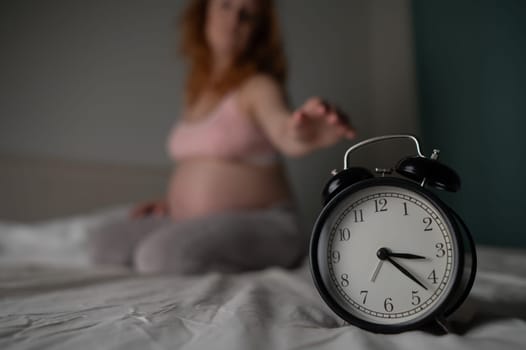 Caucasian pregnant woman sits on the bed and suffers from insomnia. Alarm clock in the foreground