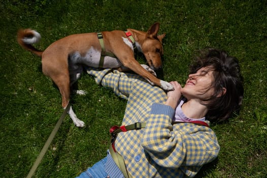 Top view of beautiful young woman and african basenji dog lying on green grass