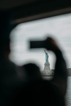 A tourist capturing the iconic Statue of Liberty from a ferry boat in New York City on a cloudy day