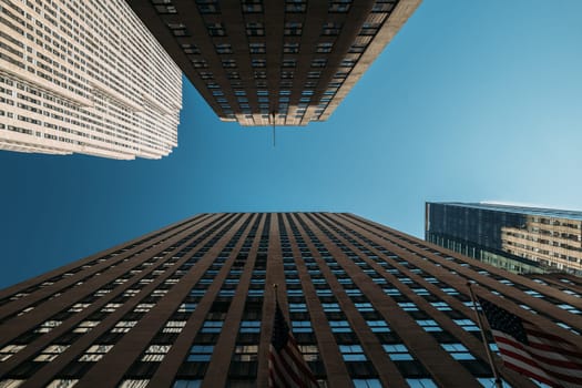 Upward view of skyscrapers in New York City against a clear blue sky. American flags are visible, emphasizing the urban architecture.