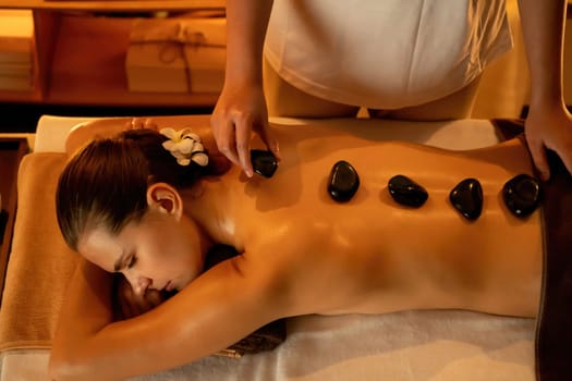 Hot stone massage at spa salon in luxury resort with warm candle light, blissful woman customer enjoying spa basalt stone massage glide over body with soothing warmth. Quiescent