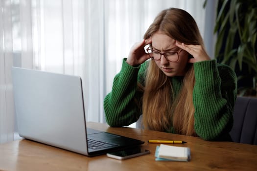 A woman wearing a green sweater is seen seated at a desk, working on a laptop computer
